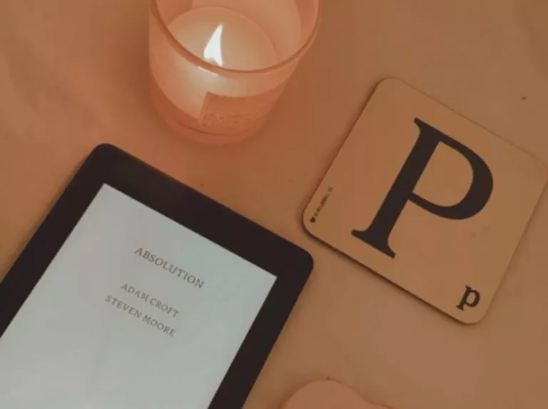 Photo shows kindle with text on screen which says Absolution. There is a square with the letter P on placed next to the kindle and a candle is lit