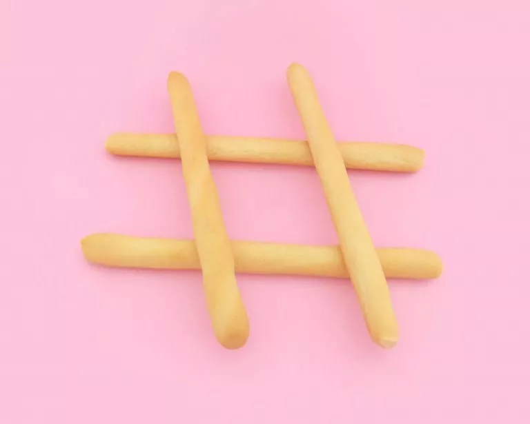 Four small brown wooden sticks creating a hashtag shape. This is on a baby pink backgroun.